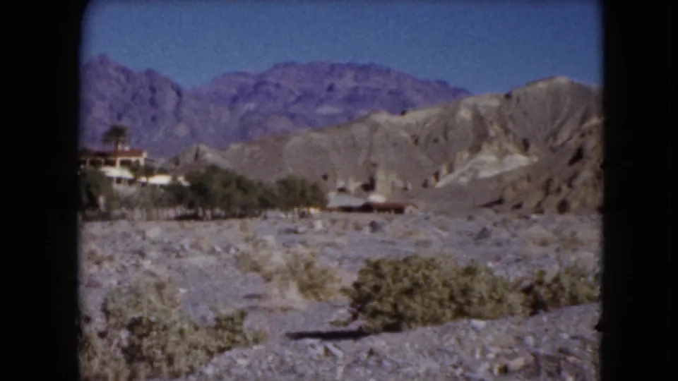 1961 Nevada 8mm frame showing desert scrub and rocky hills, with small distant buildings or roadside structures near the base of mountains.
