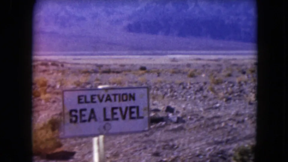 1961 Nevada 8mm frame centered on a white ELEVATION SEA LEVEL sign in sparse desert scrub with mountains beyond.
