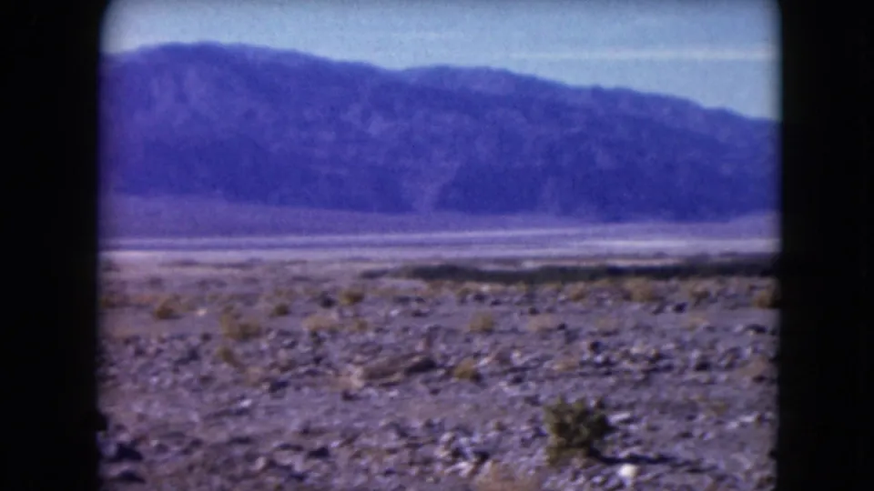 1961 Nevada 8mm frame showing a broad gravelly desert plain, sparse shrubs, and distant purple-toned mountains.