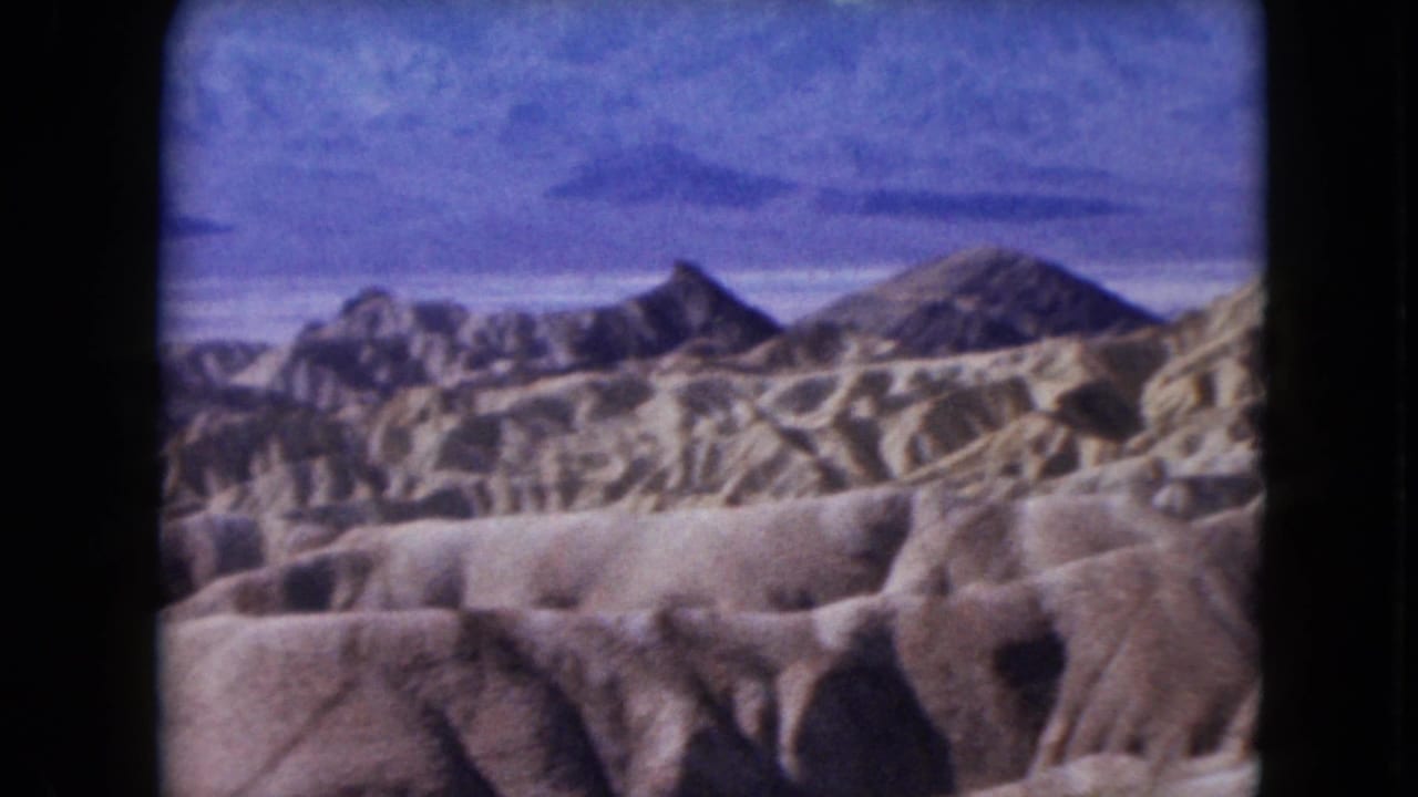 1961 Nevada 8mm frame showing tan eroded desert ridges, brown rock hills, and hazy blue-purple mountains in the distance.
