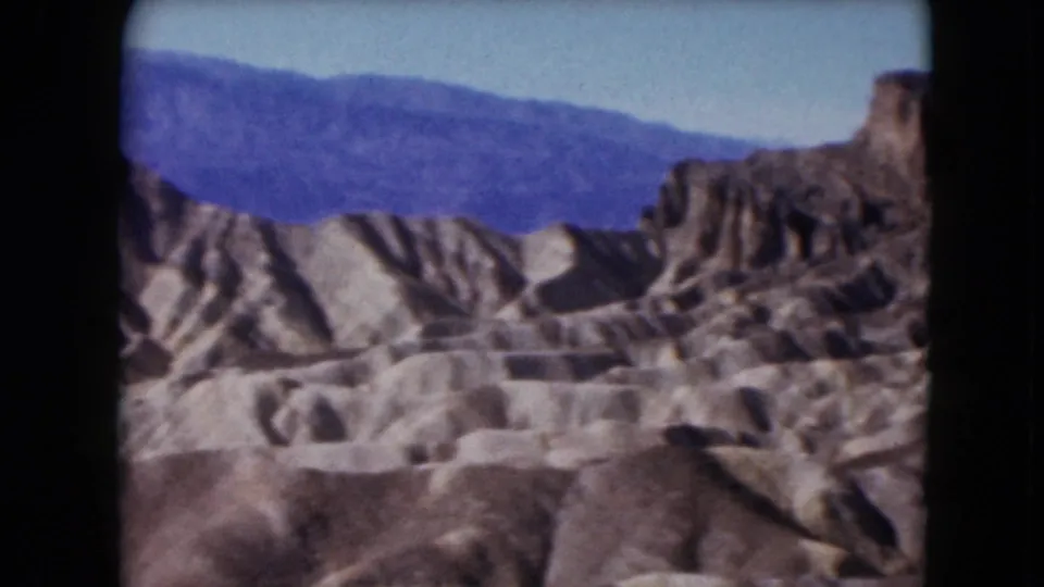 1961 Nevada 8mm frame showing dry gullies, tan ridges, and a hazy mountain backdrop in a desert landscape.
