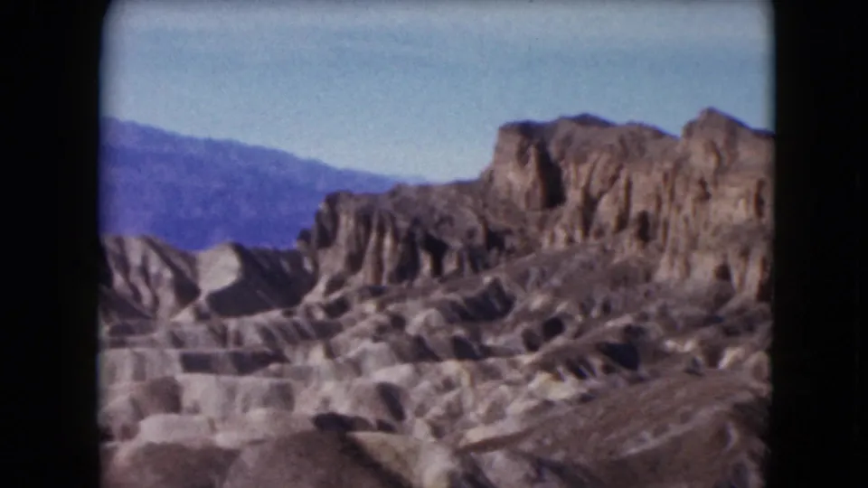 1961 Nevada 8mm frame showing a rugged rock wall, eroded foreground terrain, and hazy mountains under blue sky.