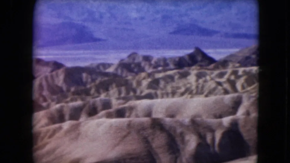 1961 Nevada 8mm frame showing rounded badlands in the foreground and darker mountain peaks beyond under a blue sky.