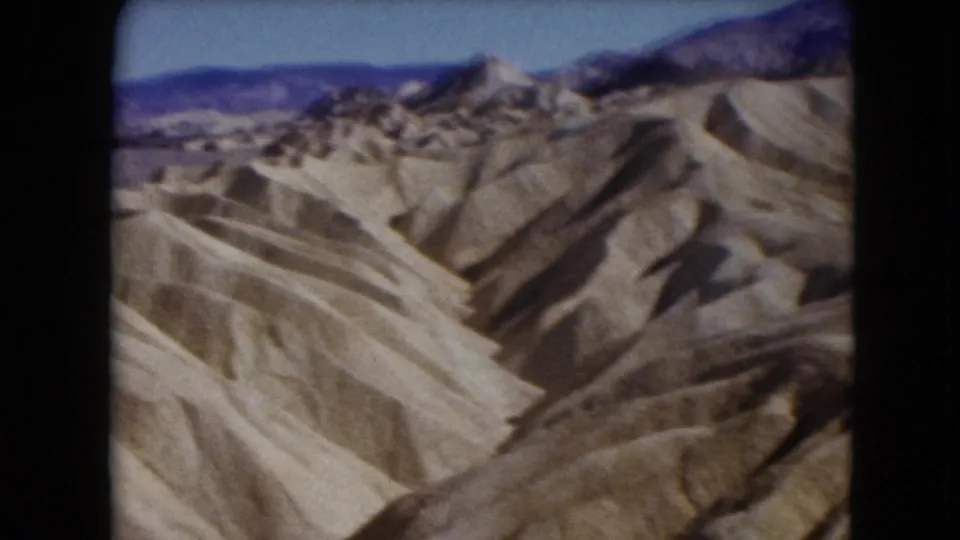 1961 Nevada home-movie frame of arid, plantless badlands stretching toward blue-purple mountains in the distance.