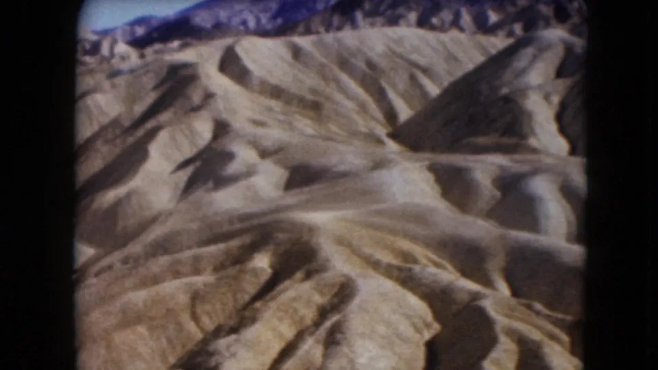 1961 Nevada 8mm frame showing dry, sculpted desert hills and gullies with darker mountains beyond.