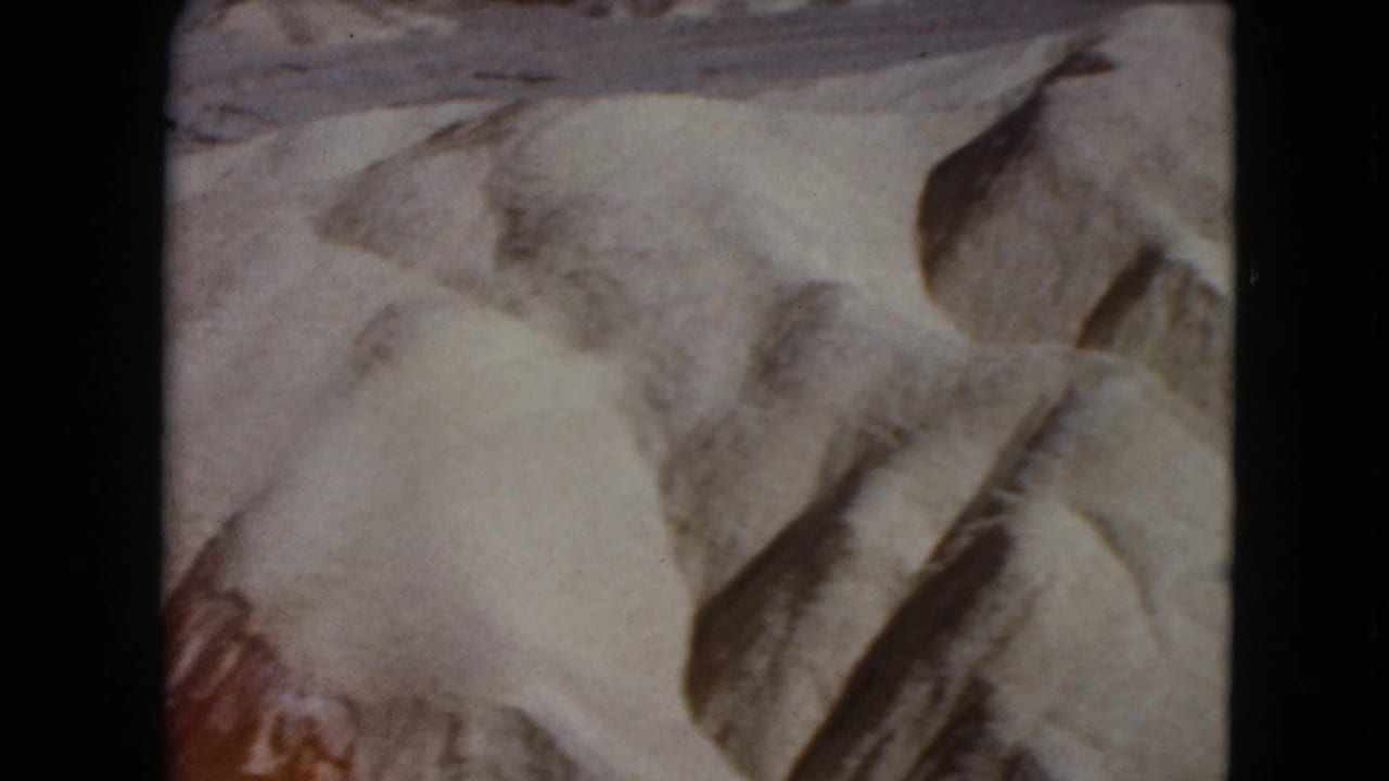 1961 Nevada 8mm frame showing pale desert sand, darker dune edges, and faint track patterns across the surface.