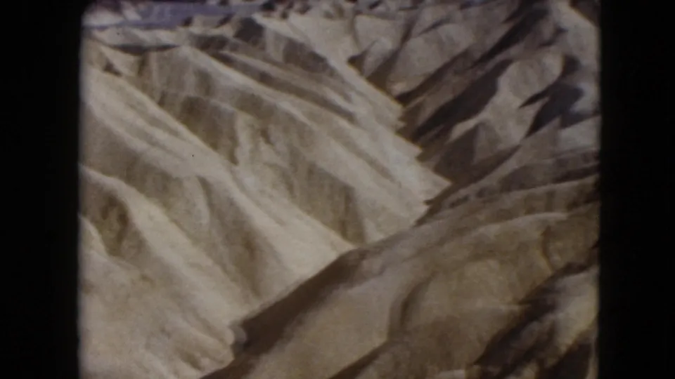 1961 Nevada desert frame showing textured dunes, crossing ridges, faint tracks, and dark shadowed slopes in 8mm footage.