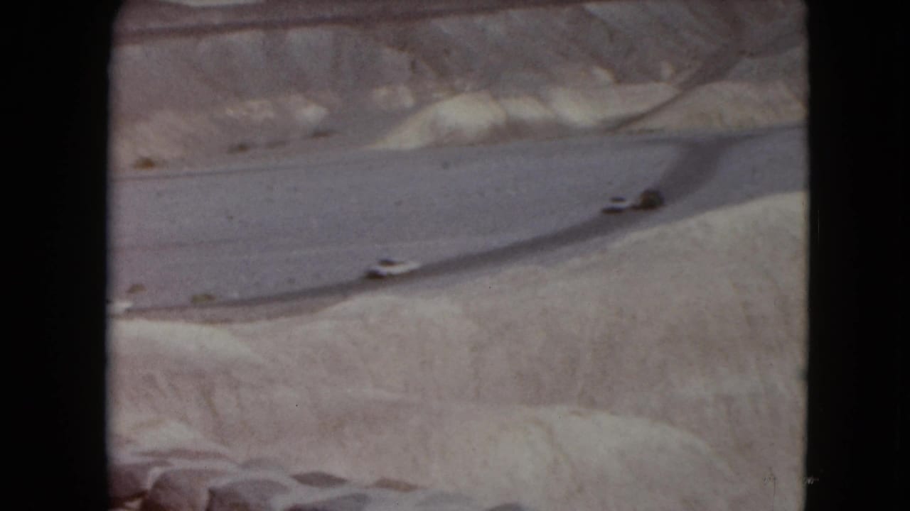 1961 Nevada home-movie frame showing distant cars on a curving road through sandy desert terrain and low rock hills.