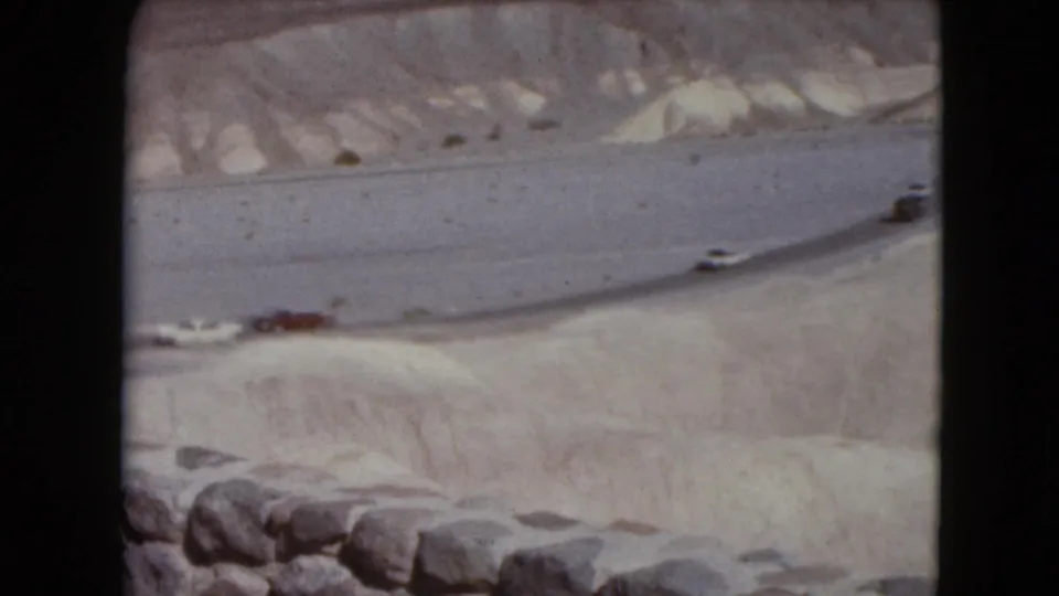 1961 8mm landscape frame showing a winding desert road below pale ridges, with small vintage cars moving through the scene.