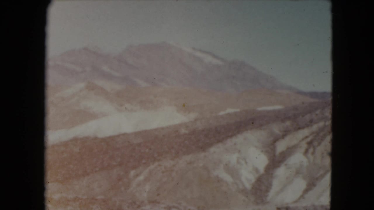 Snow-dusted mountain range above reddish rocky valley slopes under a pale sky.