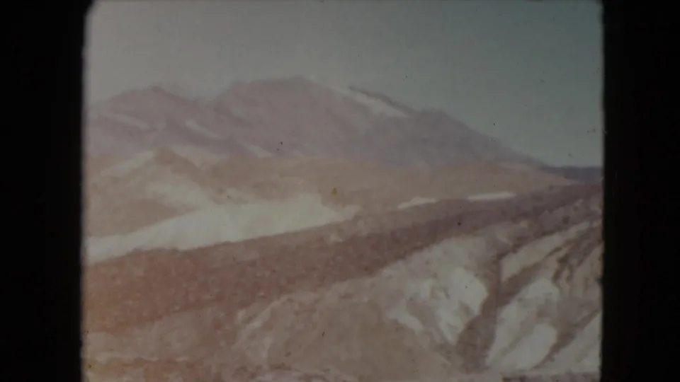 1961 Nevada 8mm frame showing a distant snow-dusted mountain range and rocky valley slopes beneath a pale sky.