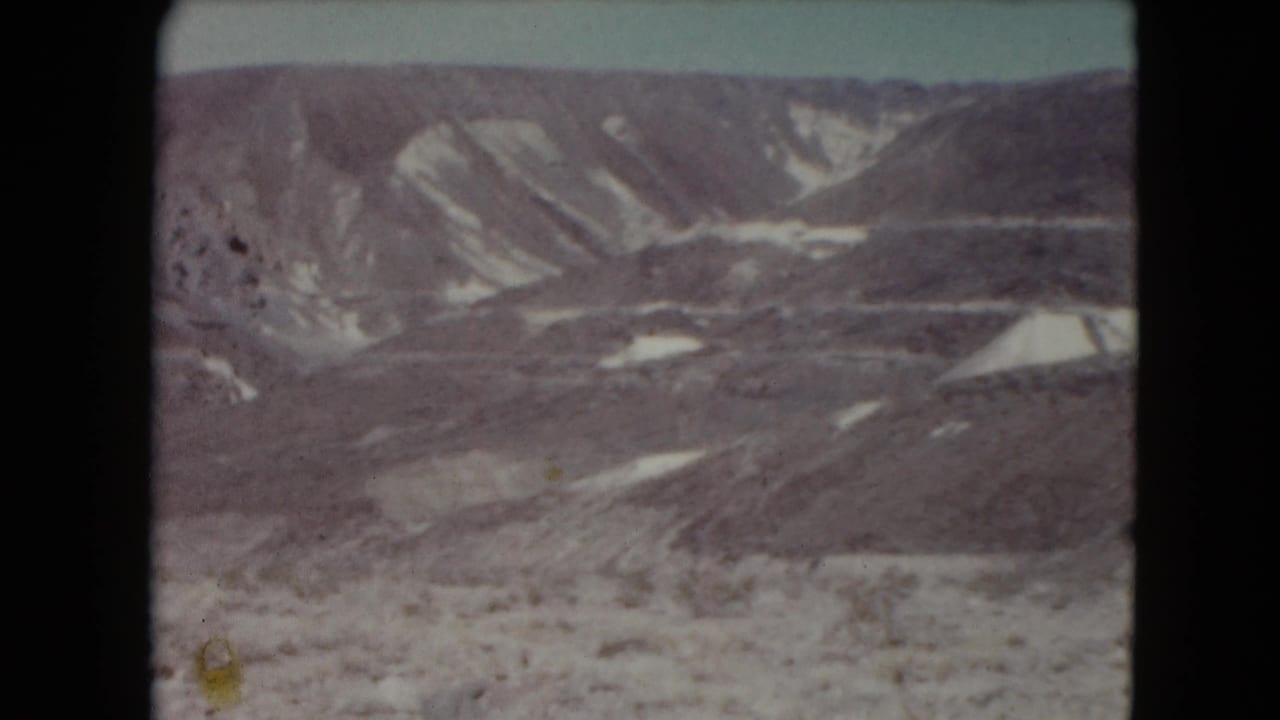 Wide desert mountain view with pale sky, scrubby foreground, and light erosion streaks.