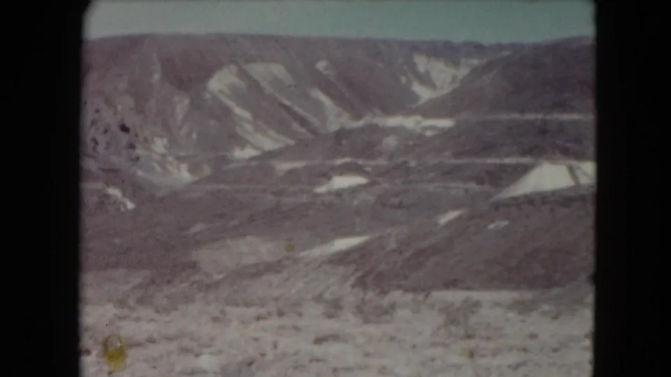 Remote Nevada desert landscape from 1961 8mm footage, showing layered hills, pale sky, and light-toned erosion marks across the slopes.