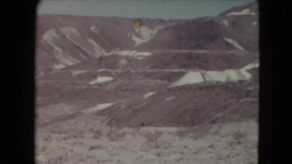 1961 Nevada home-movie frame showing dry mountain slopes, light sediment patches, and sparse foreground scrub in a remote desert setting.