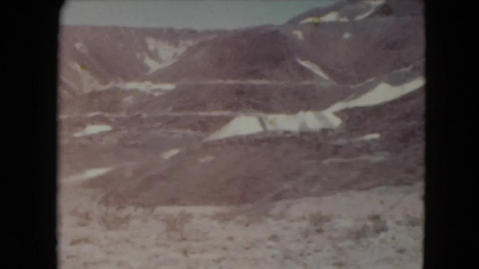 Faded 1961 8mm view of Nevada desert hills with pale sediment bands, dry foreground vegetation, and no visible human presence.