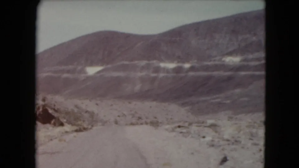 1961 Nevada 8mm frame showing a pale unpaved road, arid ground, and broad striated mountains under a muted sky.