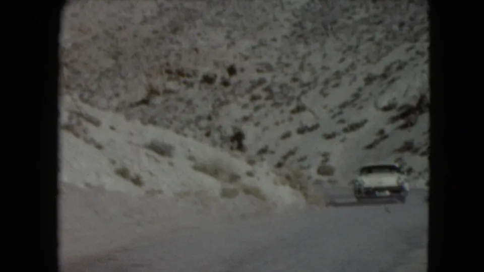 A white mid-century sedan appears at a distance on a Nevada mountain road bordered by snow-dusted rocky hills.
