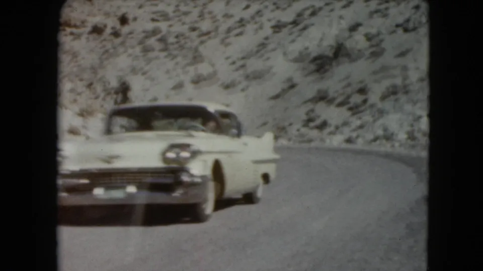 The white mid-century American sedan passes near the camera on a curving Nevada road with snowy rocky terrain behind it.
