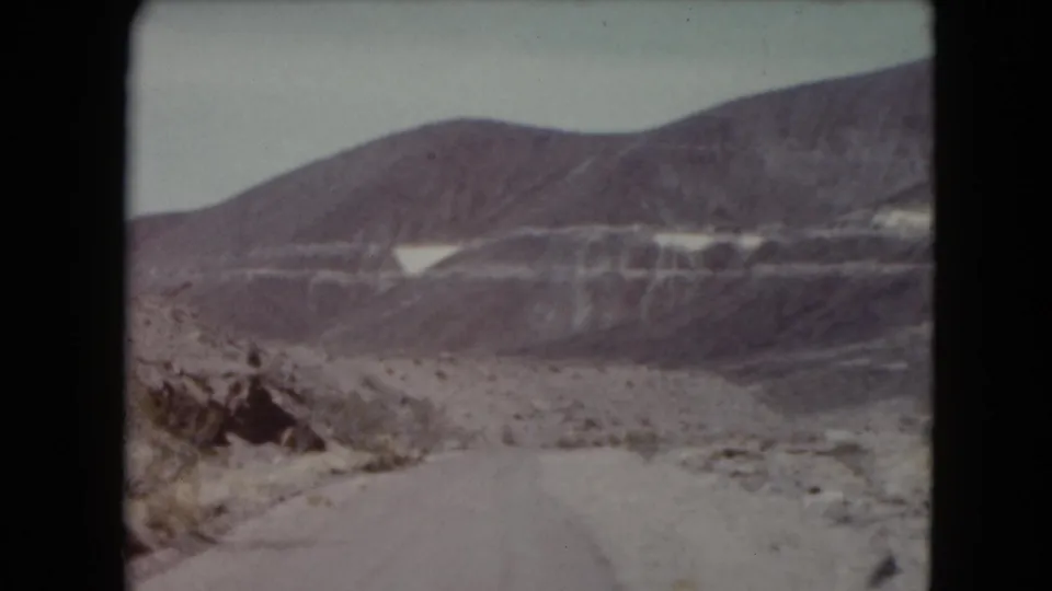 A Nevada road continues through an arid mountain landscape with pale hills, sparse brush, and distant ridges under a washed sky.