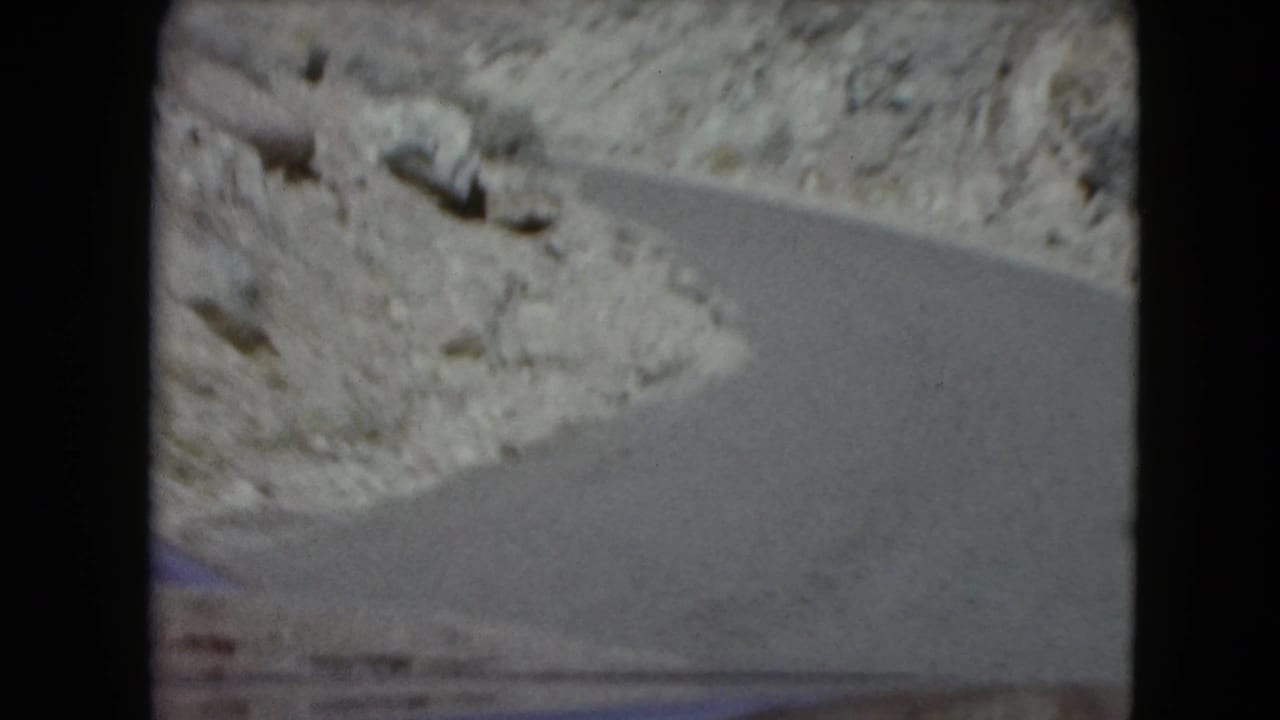 Curving paved road through rocky desert hills, seen from a moving vehicle.