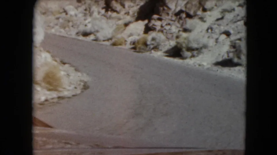 Inside-car 8mm view in Nevada as the vehicle rounds a narrow paved curve beside rocks and desert brush.