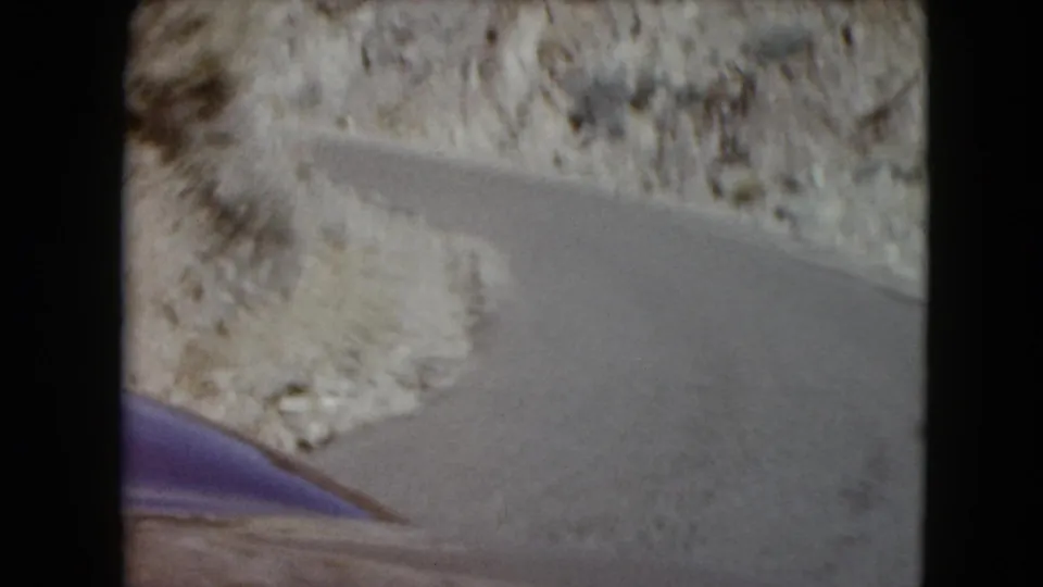 1961 Nevada home-movie frame showing the car completing a curve on a narrow road through rocky desert hills.
