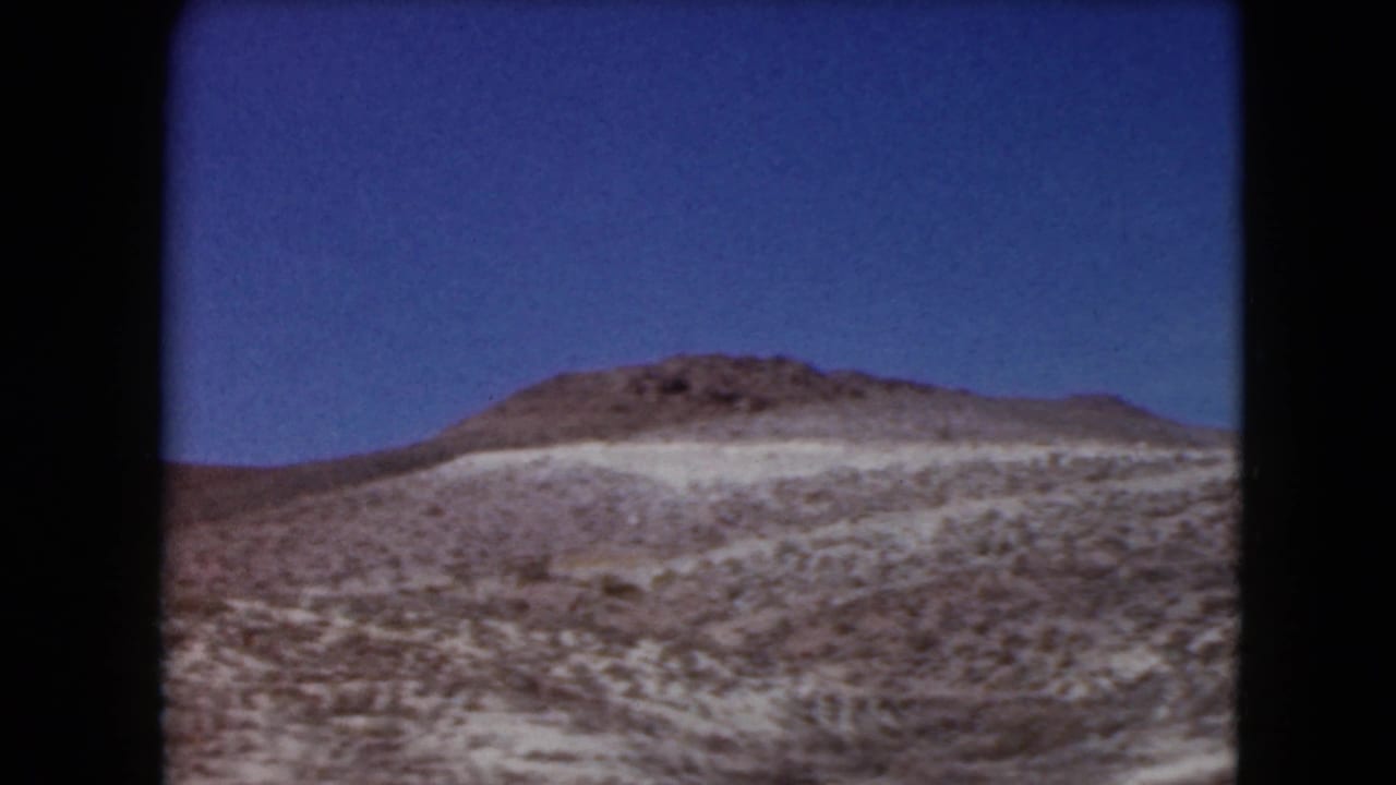 1961 Nevada home-movie frame showing an arid hill, light soil bands, and scattered scrub vegetation.