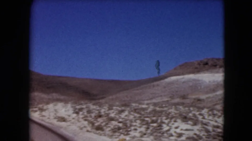 1961 Nevada 8mm frame of scrub-covered desert terrain and a low rounded hill from a traveler viewpoint.