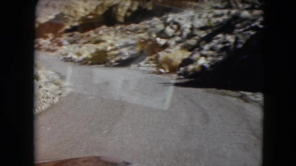 1961 Nevada 8mm frame showing a curved roadside surface beside rocky, sunlit desert terrain.