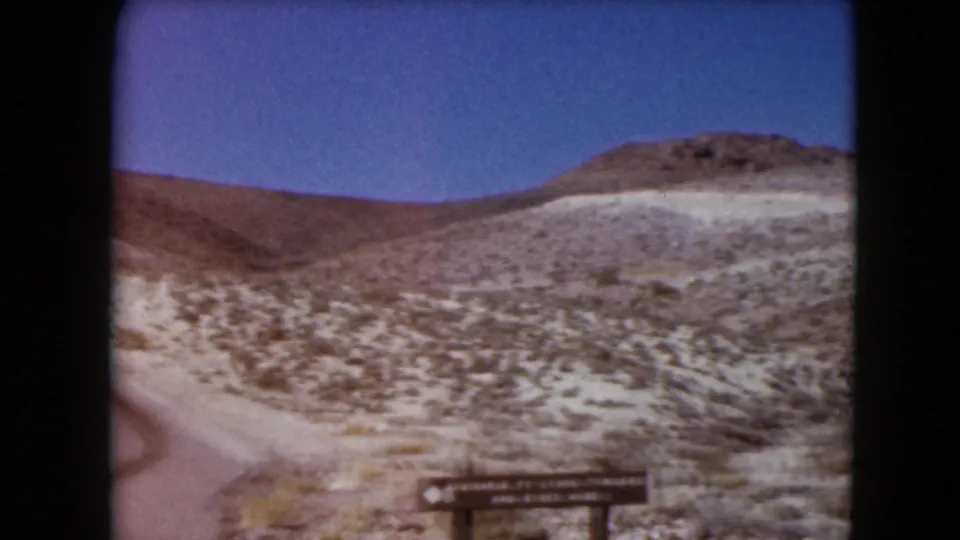 1961 Nevada travel frame showing the edge of a winding road, sparse desert vegetation, a low hill, and part of a wooden sign.