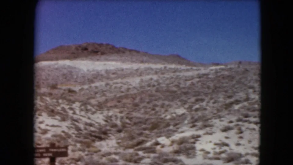 1961 Nevada 8mm frame looking across open desert scrub toward low hills, with a partial roadside sign at the lower edge.