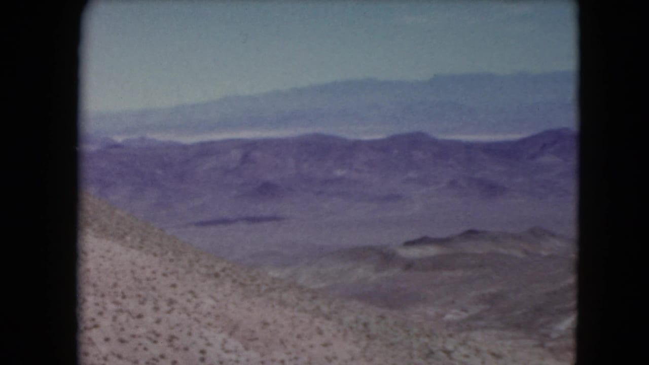 1961 Nevada home-movie frame with a scrubby desert hillside descending toward distant purple-toned mountain ranges.