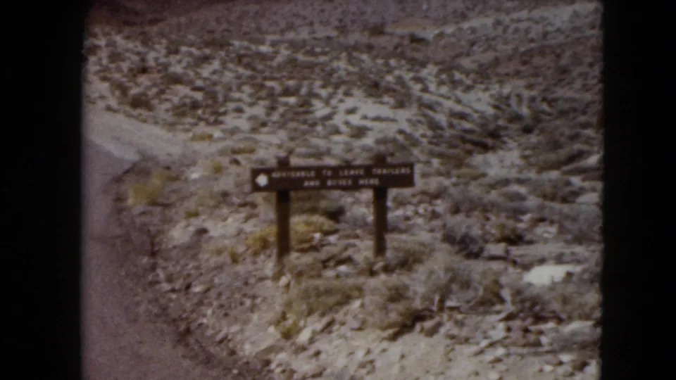 1961 Nevada 8mm frame showing a dark wooden roadside sign along a dirt road with desert scrub and mountains beyond.