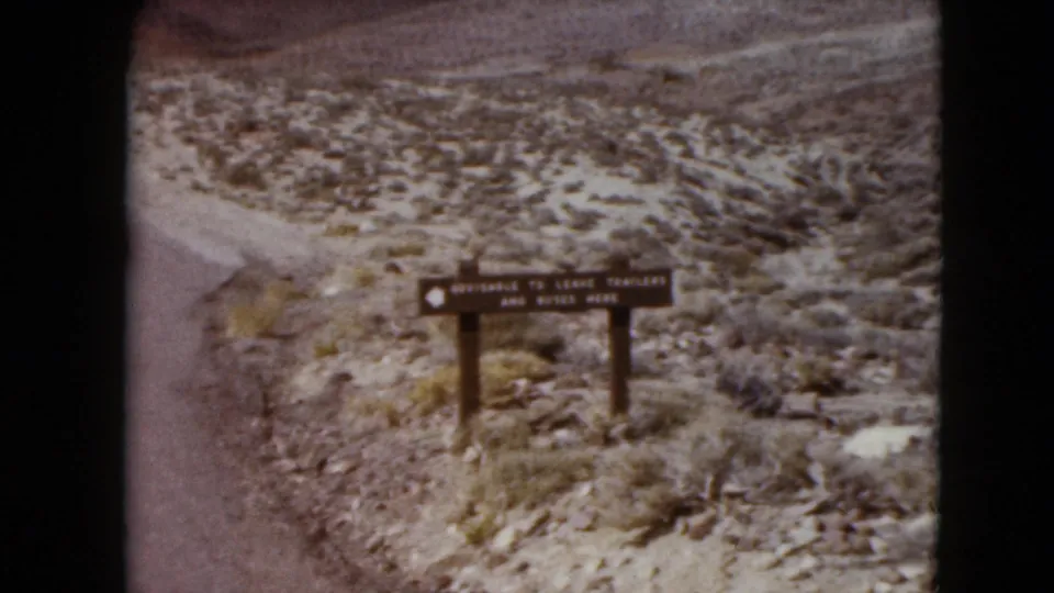 1961 Nevada home-movie frame with a wooden roadside sign, scrubby terrain, and layered desert mountains in the distance.