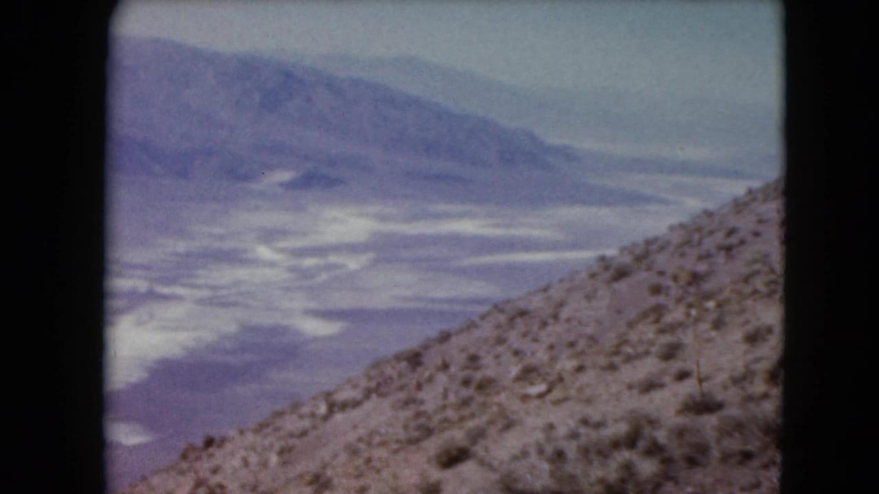 1961 Nevada travel footage frame with dry brush on a sloped foreground, pale basin below, and hazy mountain forms beyond.