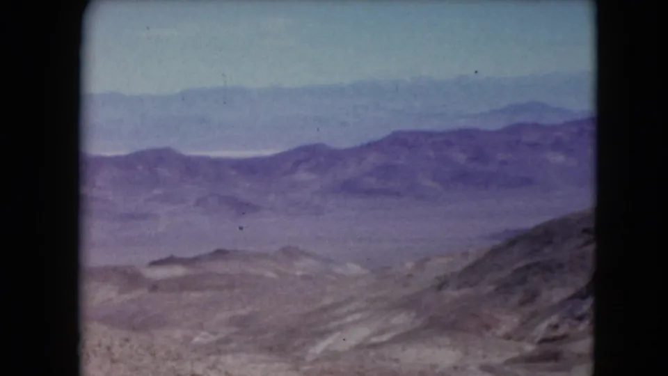 1961 Nevada home-movie frame looking across a dry valley toward low ridges and distant hazy mountain ranges.