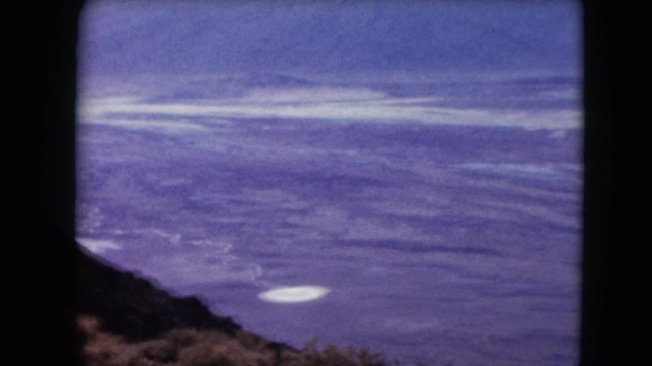 1961 8mm frame showing a high-vantage water view with rugged shoreline at lower left, a pale surface patch, and dark film borders.