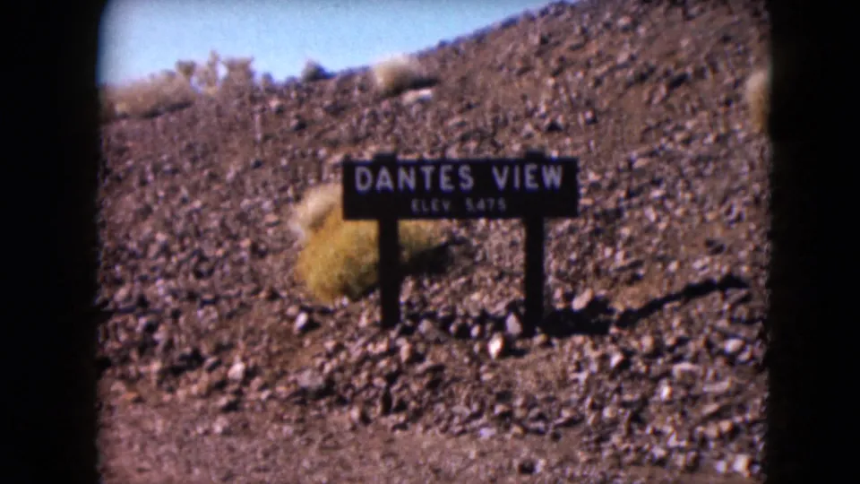 The camera holds on the Dante's View elevation sign, framed by desert rocks and sparse scrub in Death Valley.