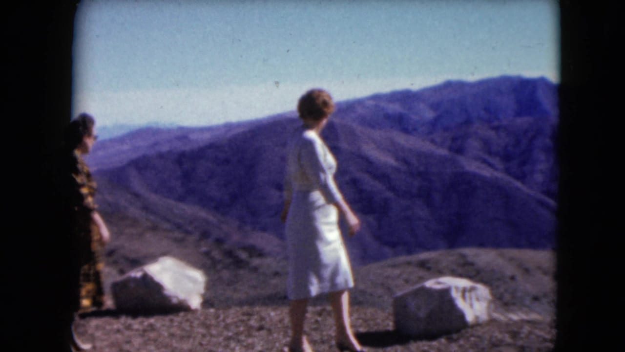 Two adult women in vintage dresses stand on a Nevada rocky overlook in 1961, facing a broad mountain landscape.