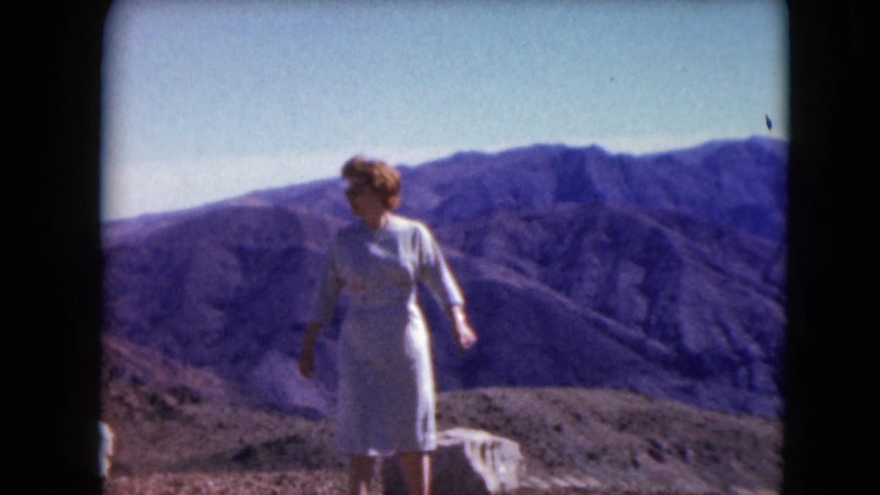 A woman in a white knee-length dress stands on a Nevada mountain overlook in 1961 with rugged hills behind her.