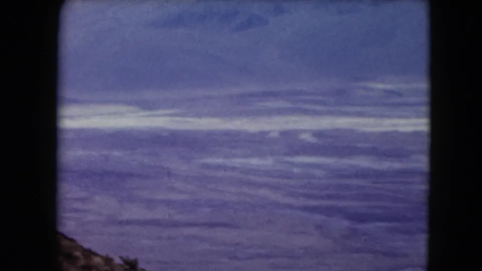 1961 Nevada home-movie frame of distant desert ridges and open sky, with a rocky slope entering the lower edge.