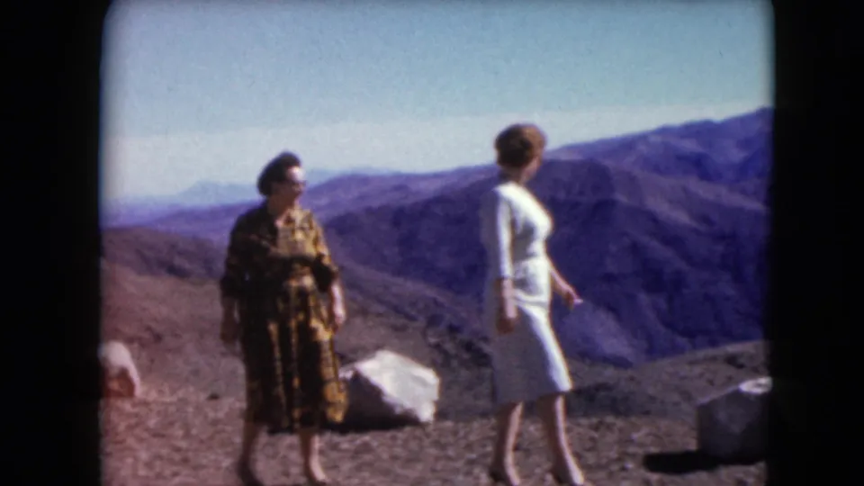 Two adult women in 1960s dress move across a Nevada desert overlook, with mountains and open sky in the background.