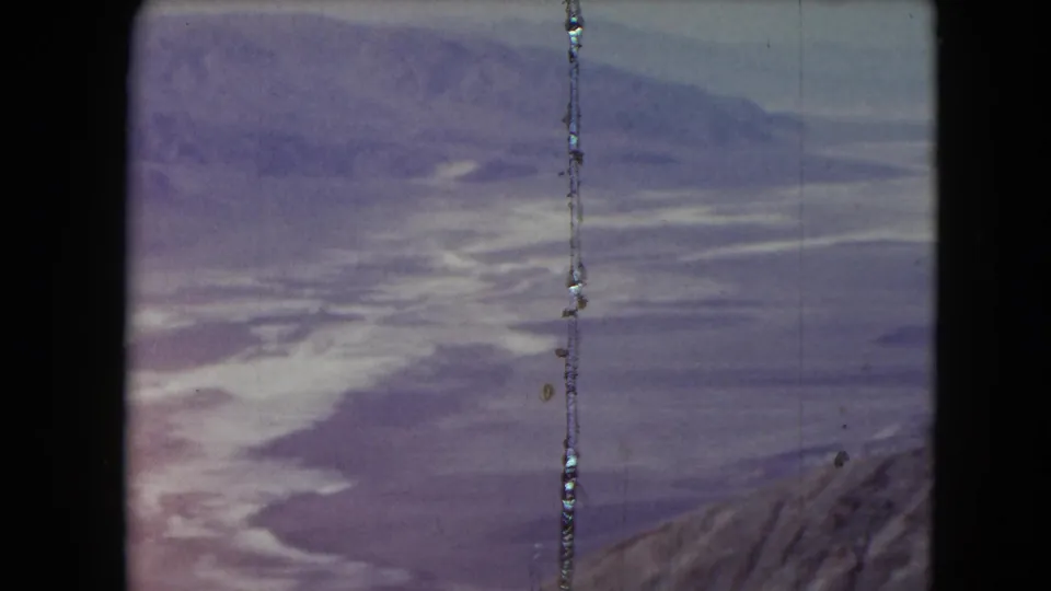 1961 Nevada 8mm frame showing soft layered mountains, pale cloud bands, a rocky lower edge, and scratched film texture.