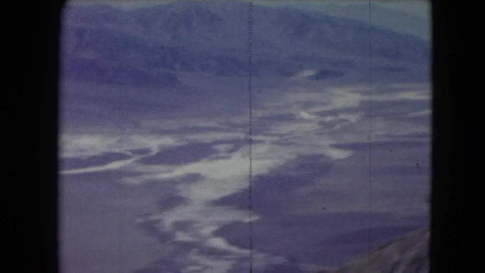 1961 Nevada 8mm frame showing distant mountains, bright white haze or cloud forms, and a small rocky edge at lower right.