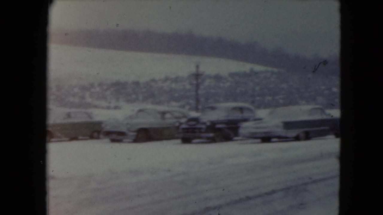 Snowy parking lot with several parked mid-century cars and hills in the background.