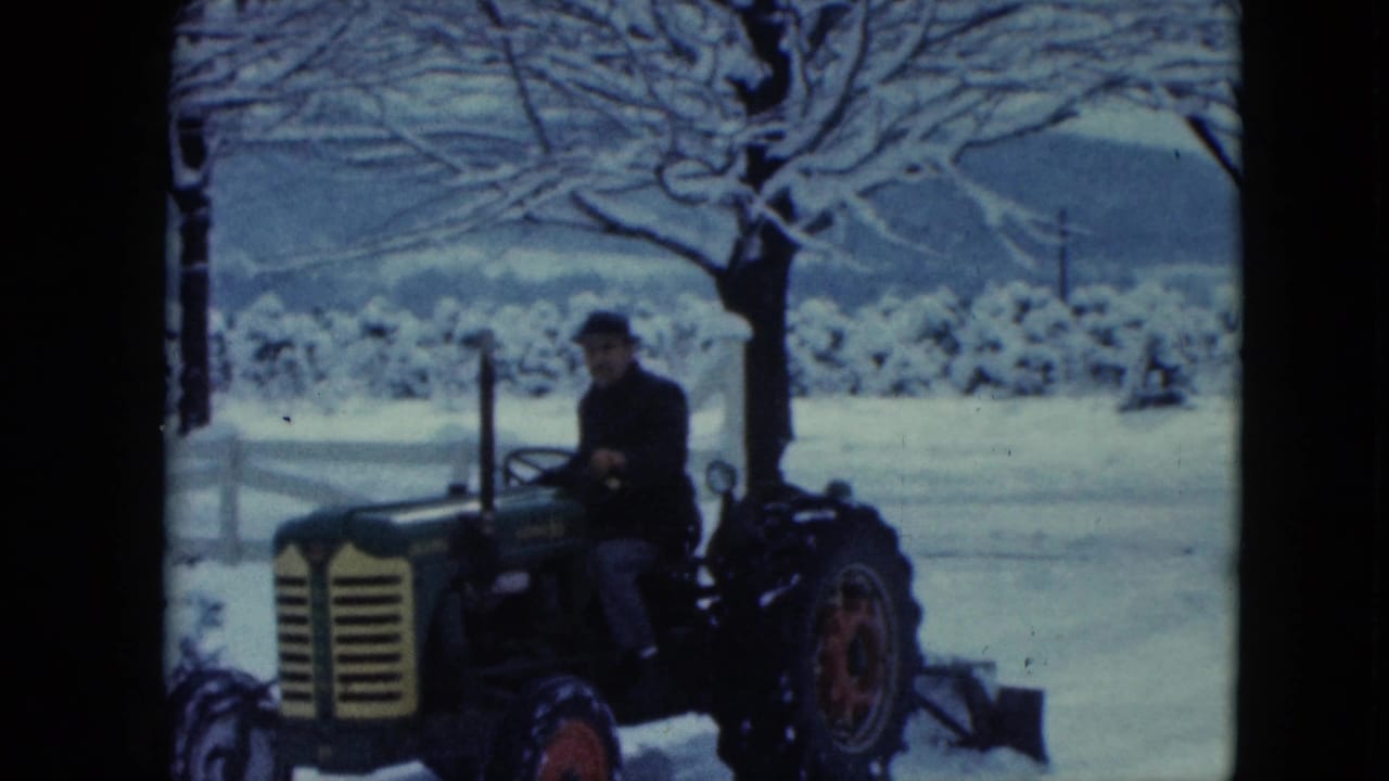 In snowy rural Nevada, an adult in dark winter clothing drives a green and yellow tractor near a white fence and bare trees.