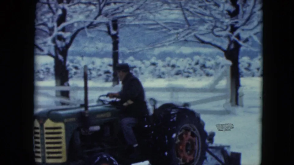An adult tractor driver passes through a snowy rural landscape with a white fence, bare trees, and distant hills visible behind.