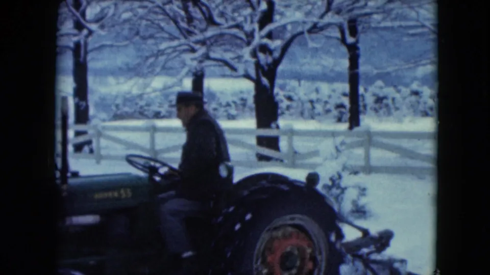 A closer side view shows the bundled driver on the tractor, with snowy branches, a white fence, and pale hills in the background.
