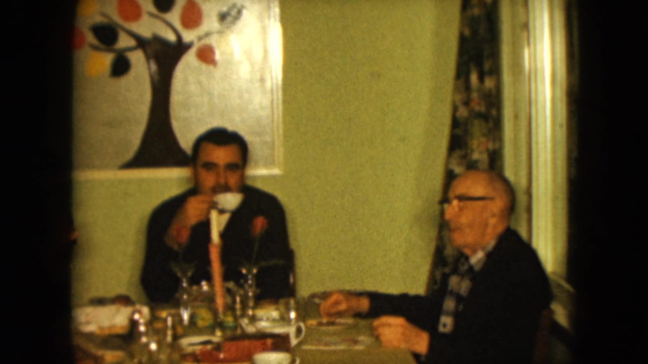 1961 Nevada home-movie frame showing two men at a dining table with glassware, a candle, and colorful tree wall art.