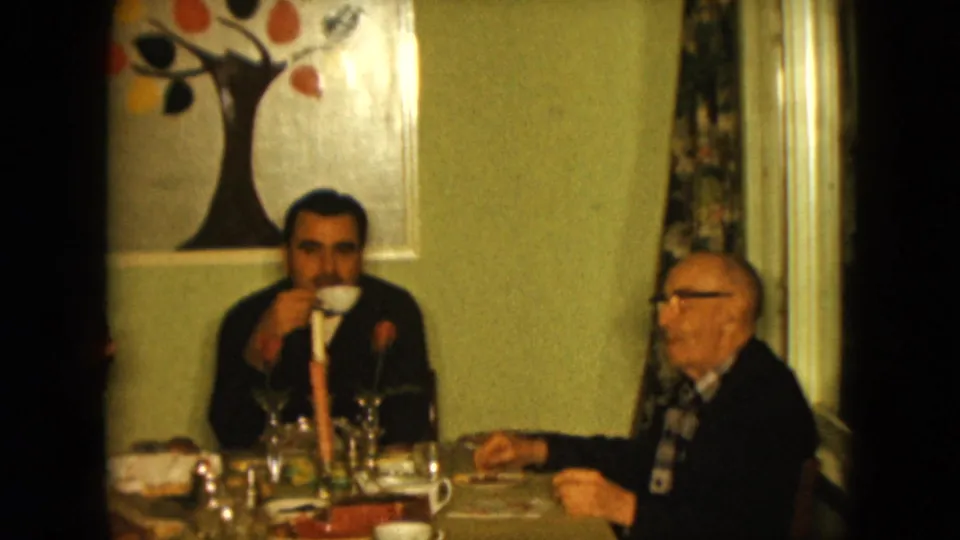 1961 Nevada home-movie frame showing two men at a dining table with glassware, a candle, and colorful tree wall art.